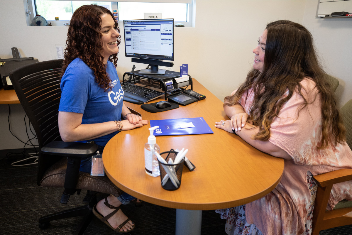 Two women sit across from each other at a desk in an office, smiling and engaged in conversation; one wears a blue Team Gesa T-shirt and the other a pink and white patterned outfit, with a computer and office supplies visible on the desk.